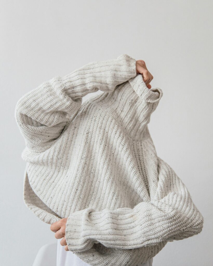 Eine Person zieht sich einen gemütlichen, beigefarbenen Strickpullover über den Kopf und verdeckt dabei das Gesicht, symbolisch für den Wunsch nach Rückzug und Komfort.